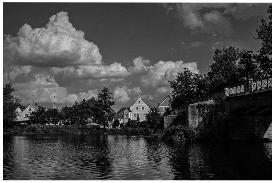Traditionelle Häuser und Brücke am Flussufer unter bewölktem Himmel, flankiert von Bäumen, © Klaus Gruber | dolphin photography