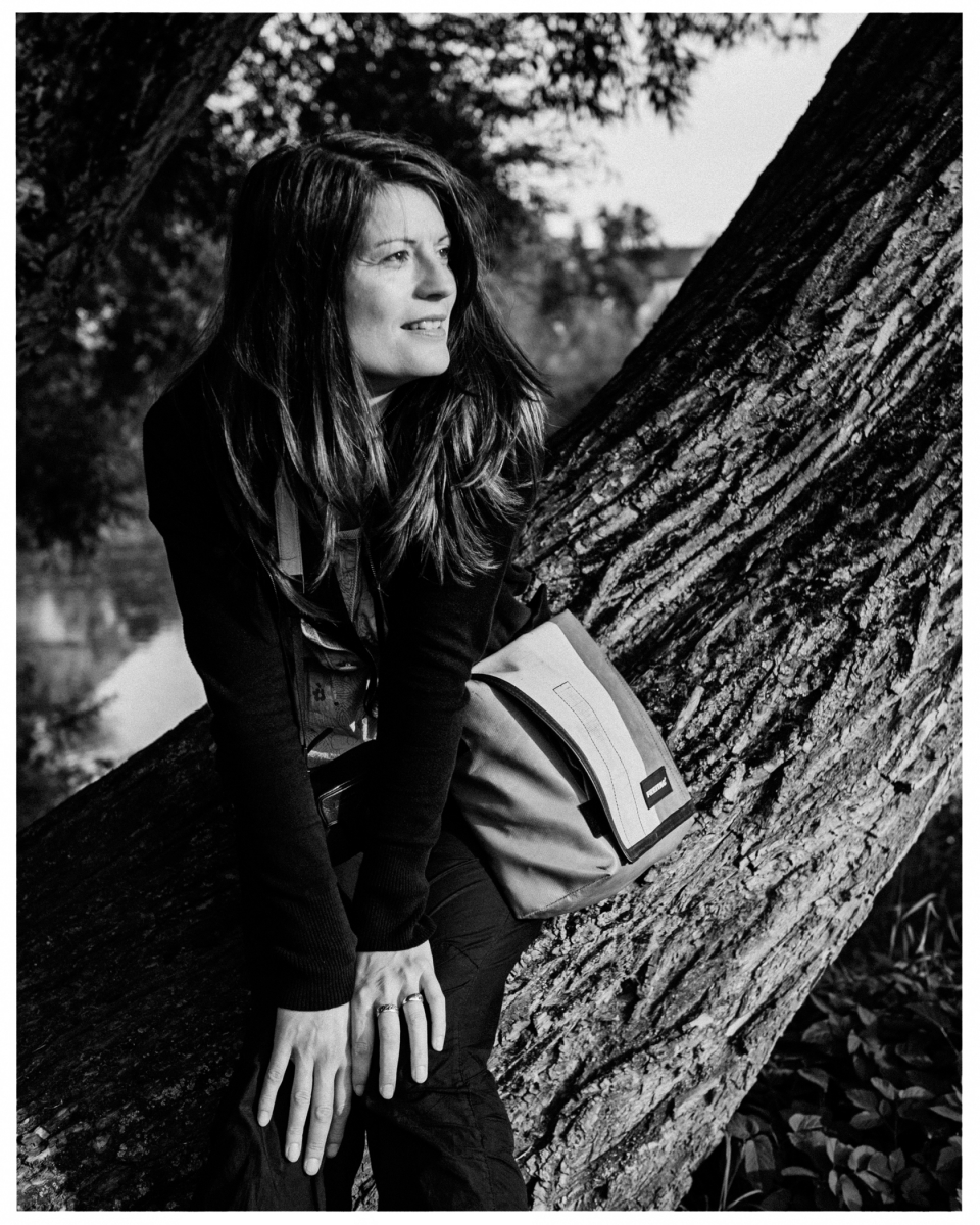 Person sitzt lächelnd auf Baumstamm in der Natur; Schwarz-Weiß-Fotografie mit Umhängetasche, © Klaus Gruber | dolphin photography