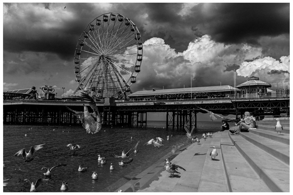 Riesenrad auf einem Pier, umgeben von Möwen und dunklen Wolken, Menschen auf Stufen sitzend, © Klaus Gruber | Fotograf Fürth