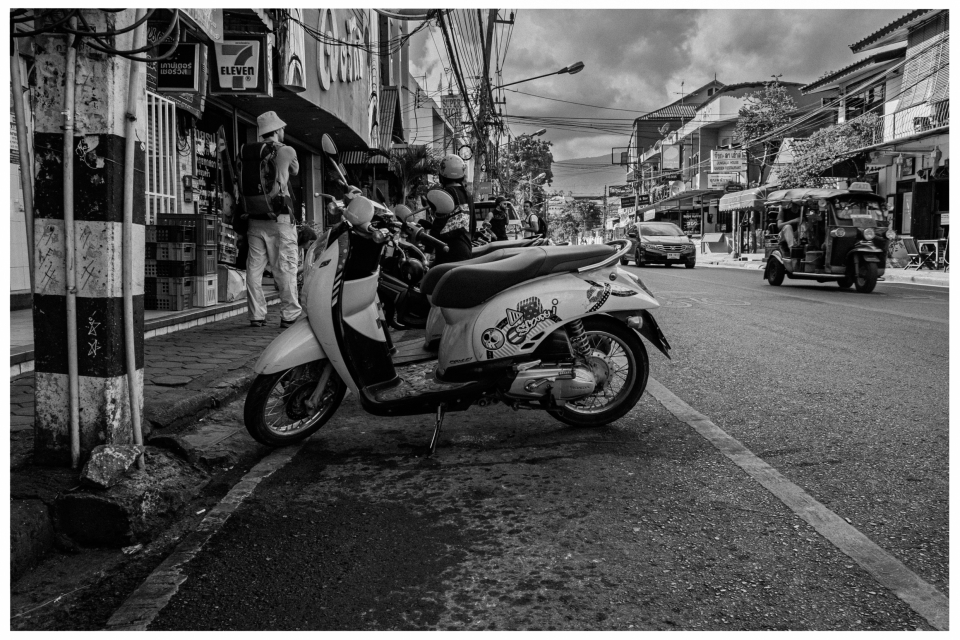 Straßenszene mit Motorrad, Personen vor Geschäft, Autos und Tuk-Tuk auf urbaner Straße, © Klaus Gruber | Fotograf Fürth