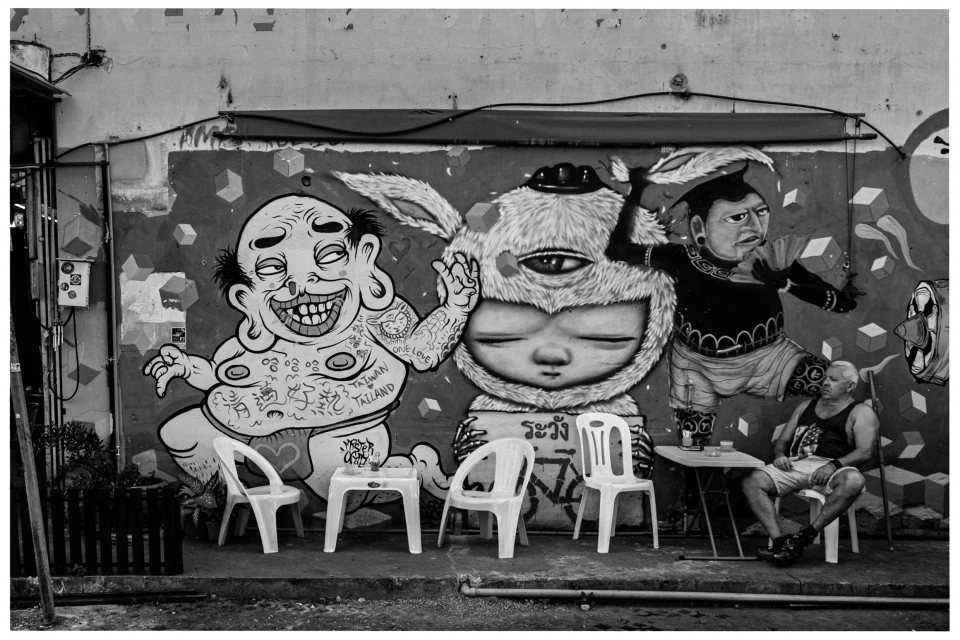 Straßenszene mit Graffiti von drei Figuren, Plastikstühlen und sitzender Person am Tisch, © Klaus Gruber | Fotograf Fürth