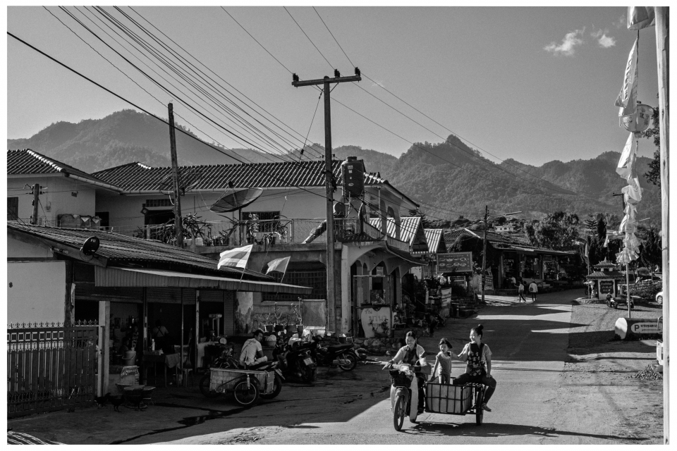 Dorfstraße mit Wohnhäusern, Geschäften, Familie auf Motorrad und Berge im Hintergrund, © Klaus Gruber | Fotograf Fürth