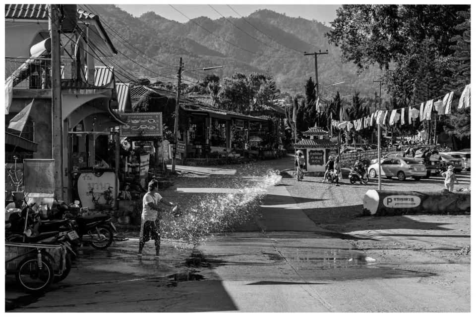 Eine Person spritzt Wasser auf eine Straße, umgeben von Gebäuden, Fahrzeugen und bewaldeten Hügeln, © Klaus Gruber | Fotograf Fürth
